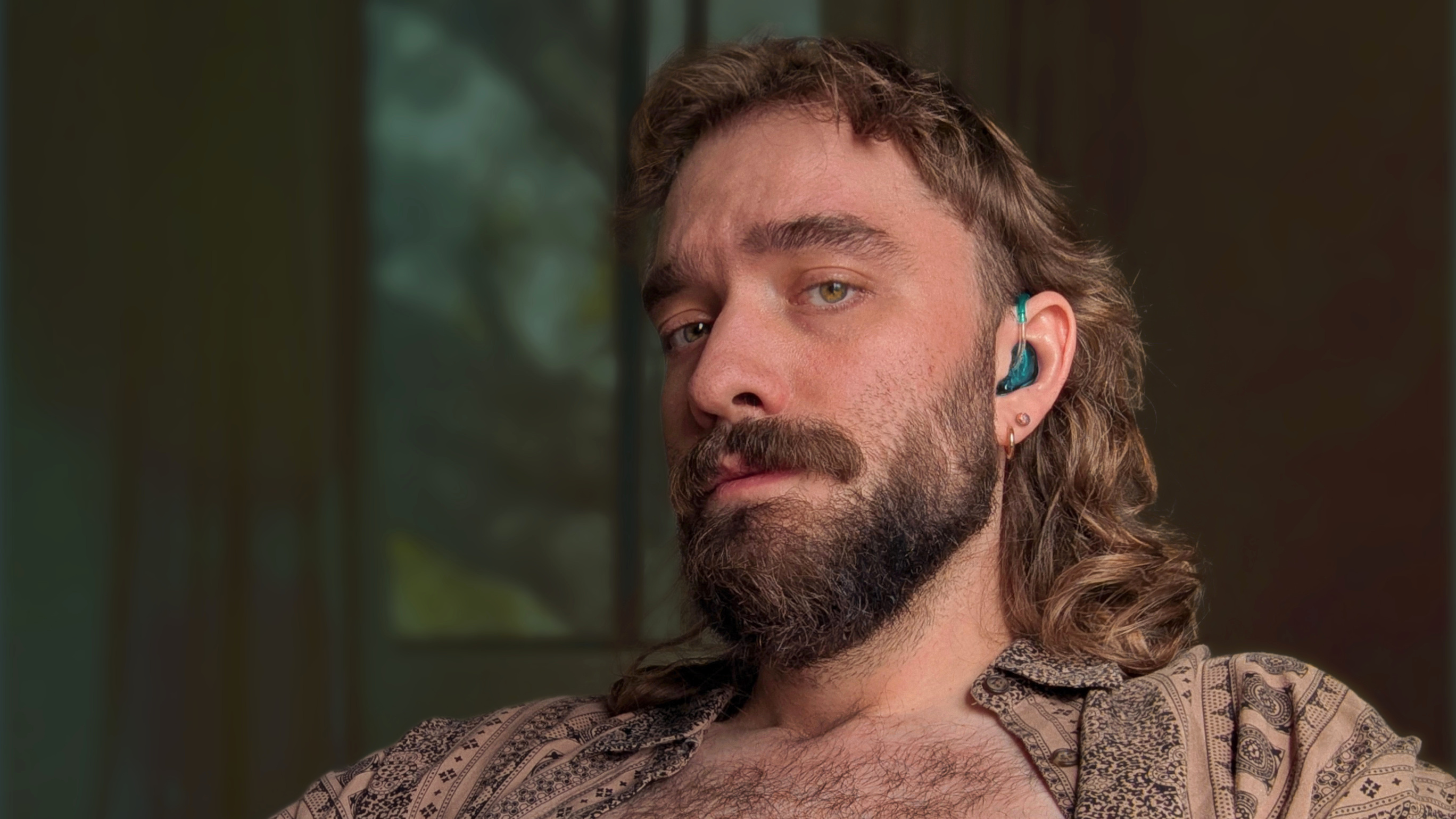 Photo of Hayden, a white man with long brown wavy hair, a scruffy beard, and a blue hearing aid, looking at the camera, with a hazy, green-and-blue-tinted room behind him.
